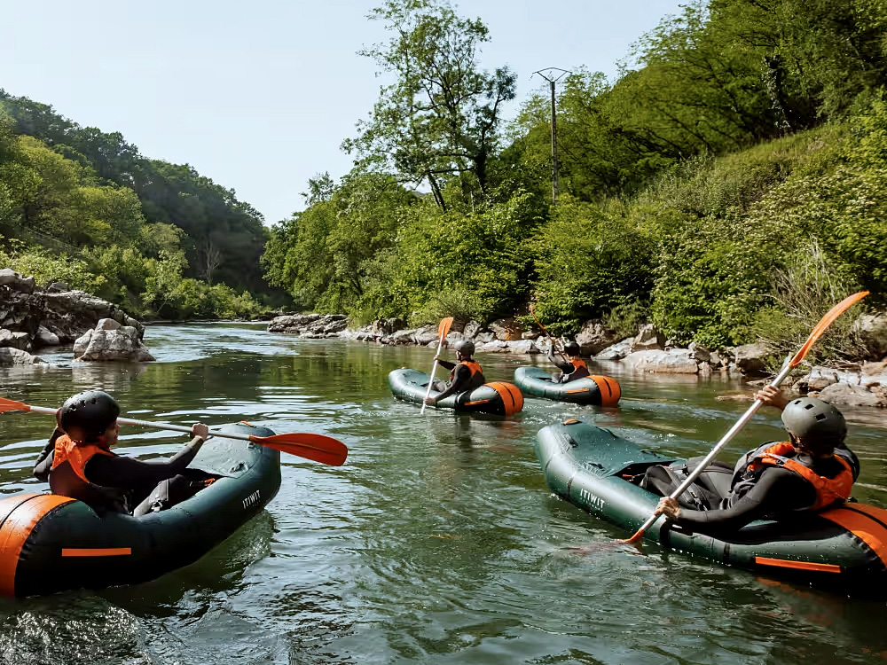 Das Bild zeigt eine Gruppe von vier Personen, die in aufblasbaren **Packrafts** auf einem ruhigen Fluss paddeln. Sie tragen **Schwimmwesten und Helme**, was auf eine sichere Outdoor-Aktivität hinweist. Die Umgebung ist geprägt von **grünen Wäldern, felsigen Ufern und klarem Wasser**, was eine idyllische und naturbelassene Atmosphäre schafft. Die Paddler bewegen sich mit **orangefarbenen Doppelpaddeln** durch das Wasser und scheinen gemeinsam auf einer entspannten oder abenteuerlichen Tour unterwegs zu sein.