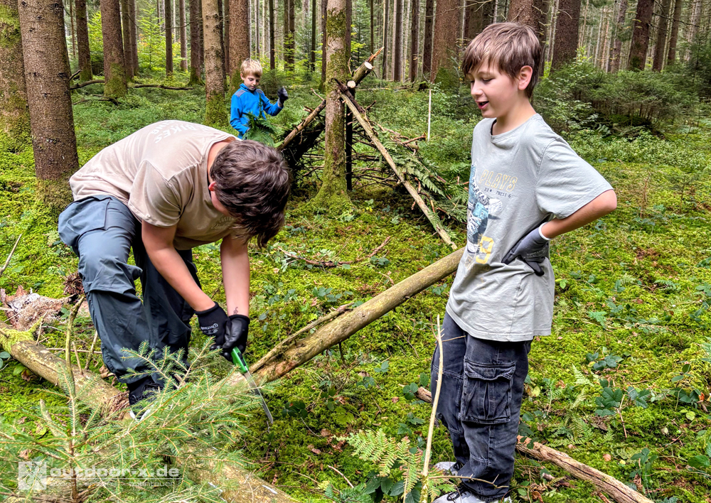Outdoor-Abenteuer für Vater und Kind: Beim gemeinsamen Shelterbau im Wald bearbeiten Vater und Sohn einen Baumstamm, während im Hintergrund ein weiteres Kind an einer Notunterkunft aus Ästen arbeitet.