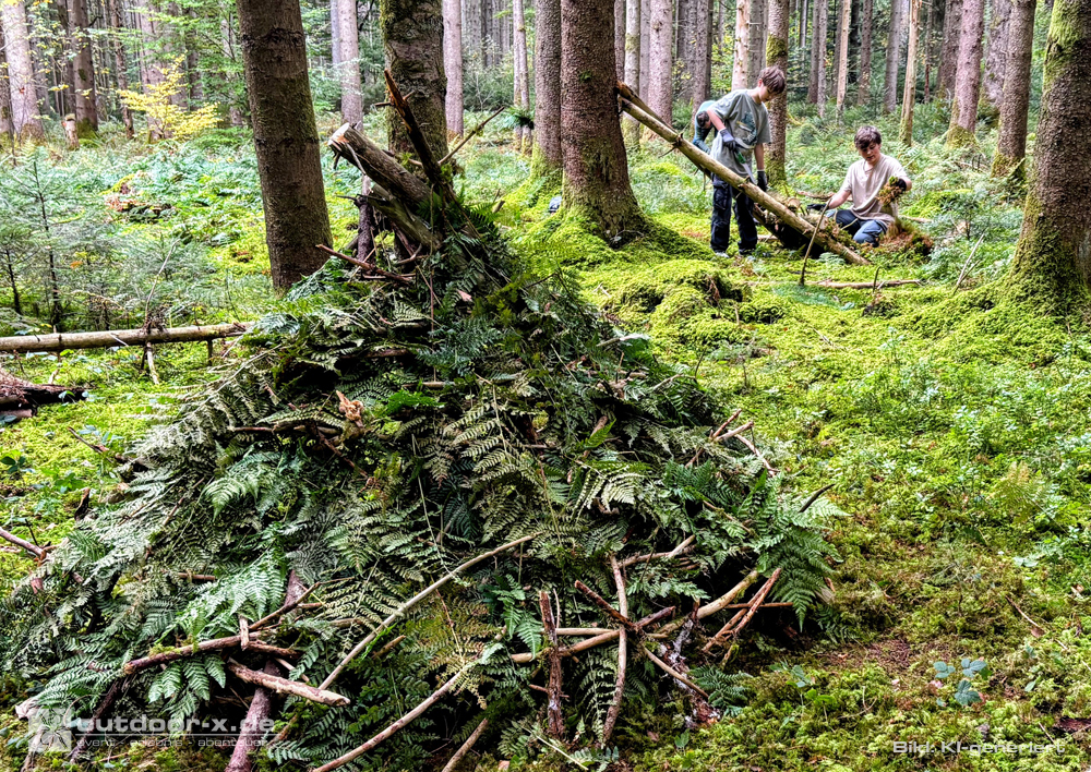 Outdoor-X Academy Outdoor-Abenteuer für Vater und Kind: Selbst gebauter Shelter aus Ästen und Farnen im Wald, während Vater und Sohn im Hintergrund gemeinsam eine weitere Notunterkunft errichten.