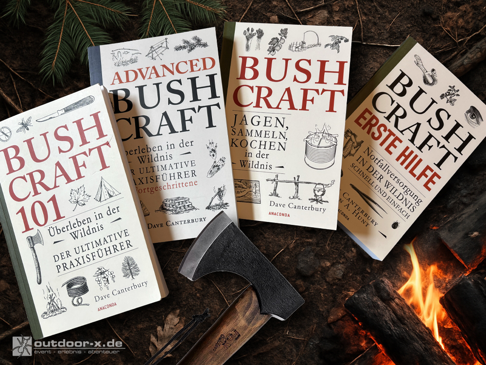 Das Bild zeigt vier auf Waldboden liegende Bücher der Bushcraft-Reihe von Dave Canterbury: „Bushcraft 101 – Überleben in der Wildnis“, „Advanced Bushcraft – Der ultimative Praxisführer für Fortgeschrittene“, „Bushcraft – Jagen, Sammeln, Kochen in der Wildnis“ und „Bushcraft Erste Hilfe – Notfallversorgung in der Wildnis“. Neben den Büchern liegt ein kleines Beil mit Holzgriff. Am unteren Bildrand ist das Logo von „outdoor-x.de“ eingeblendet. Rechts leuchtet ein kleines Lagerfeuer, während im Hintergrund Tannenzweige den Waldboden säumen.