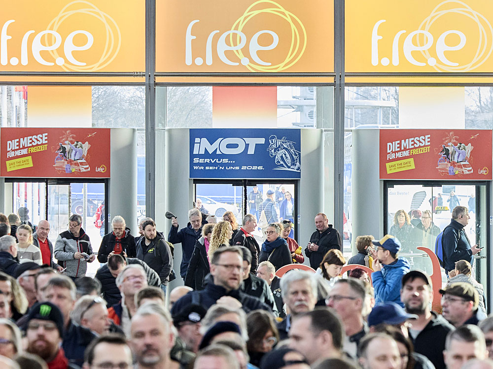 Ein geschäftiger Eingangsbereich einer Freizeit- und Reisemesse mit vielen Besuchern. Große Werbeschilder für die Messe 'f.re.e' hängen über den Türen, während Menschen in Jacken und Mützen durcheinandergehen, einige mit Handys oder Rucksäcken. Im Hintergrund sind Glastüren, durch die weitere Besucher eintreten. Plakate für die 'IMOT'-Motorradmesse und die Freizeitmesse sind über den Eingängen sichtbar.