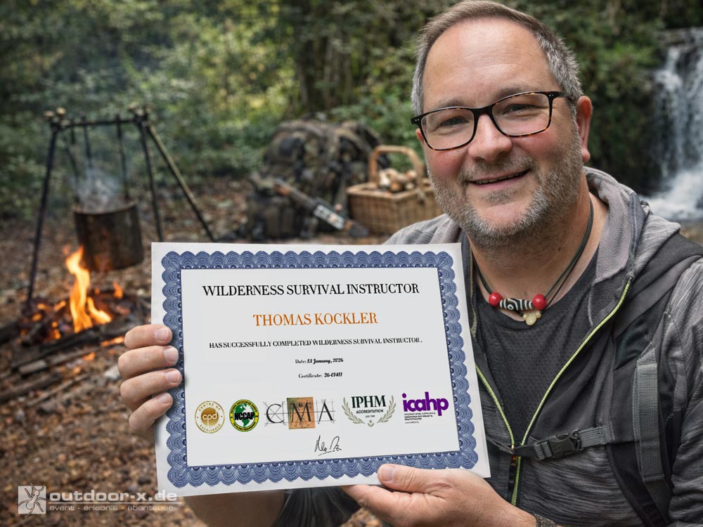 Thomas hält im Wald ein Zertifikat zum „Wilderness Survival Instructor“ in die Kamera; im Hintergrund Lagerfeuer, Outdoor-Ausrüstung und ein kleiner Wasserfall. Der Hintergrund ist KI-generiert.