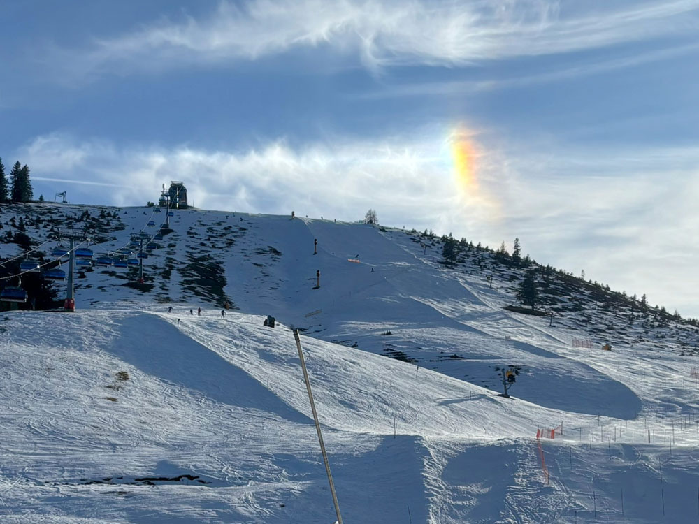 Auf dem Bild ist eine Skipiste im Skigebiet Sudelfeld zu sehen. Die Pisten sind mit Kunstschnee bedeckt, während die umliegende Landschaft größtenteils schneefrei ist und braun-grüne Flächen zeigt. Ein Sessellift führt den Hang hinauf, einige Skifahrer sind auf den Pisten unterwegs. Am Himmel ist eine auffällige, regenbogenartige Lichterscheinung (eine sogenannte Nebensonne) zu erkennen.