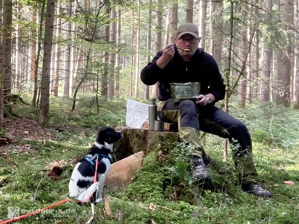 Das Bild zeigt den Redakteur, der auf einem Baumstumpf im Wald sitzt und eine Mahlzeit aus der CONVAR Feldküche probiert. Neben ihm sitzt sein Hund an einer roten Schleppleine auf dem moosbedeckten Waldboden und beobachtet ihn aufmerksam.